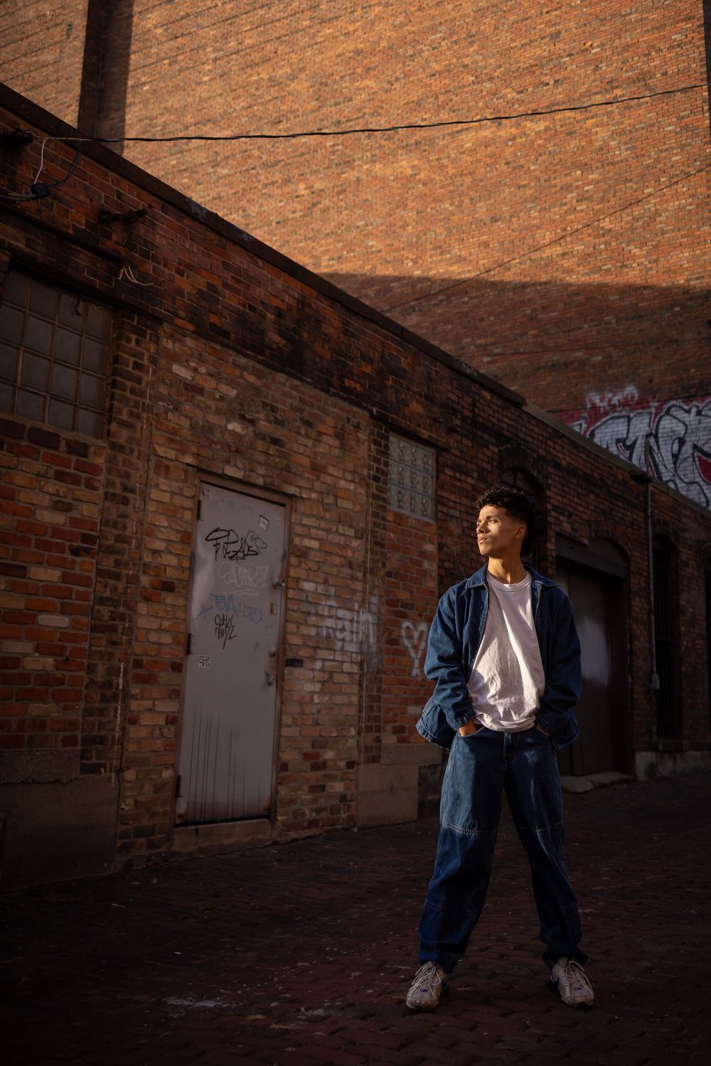 A person poses against a brick wall in casual denim clothing at dusk with dramatic lighting.
