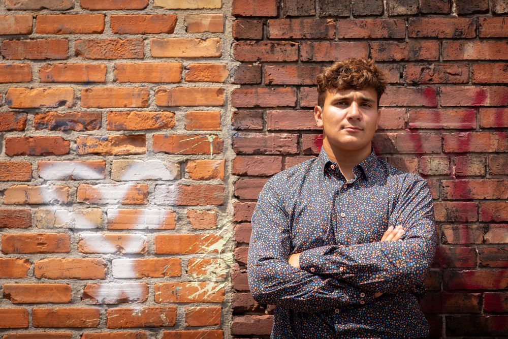 Professional headshot against a rustic brick wall background with crossed arms pose.