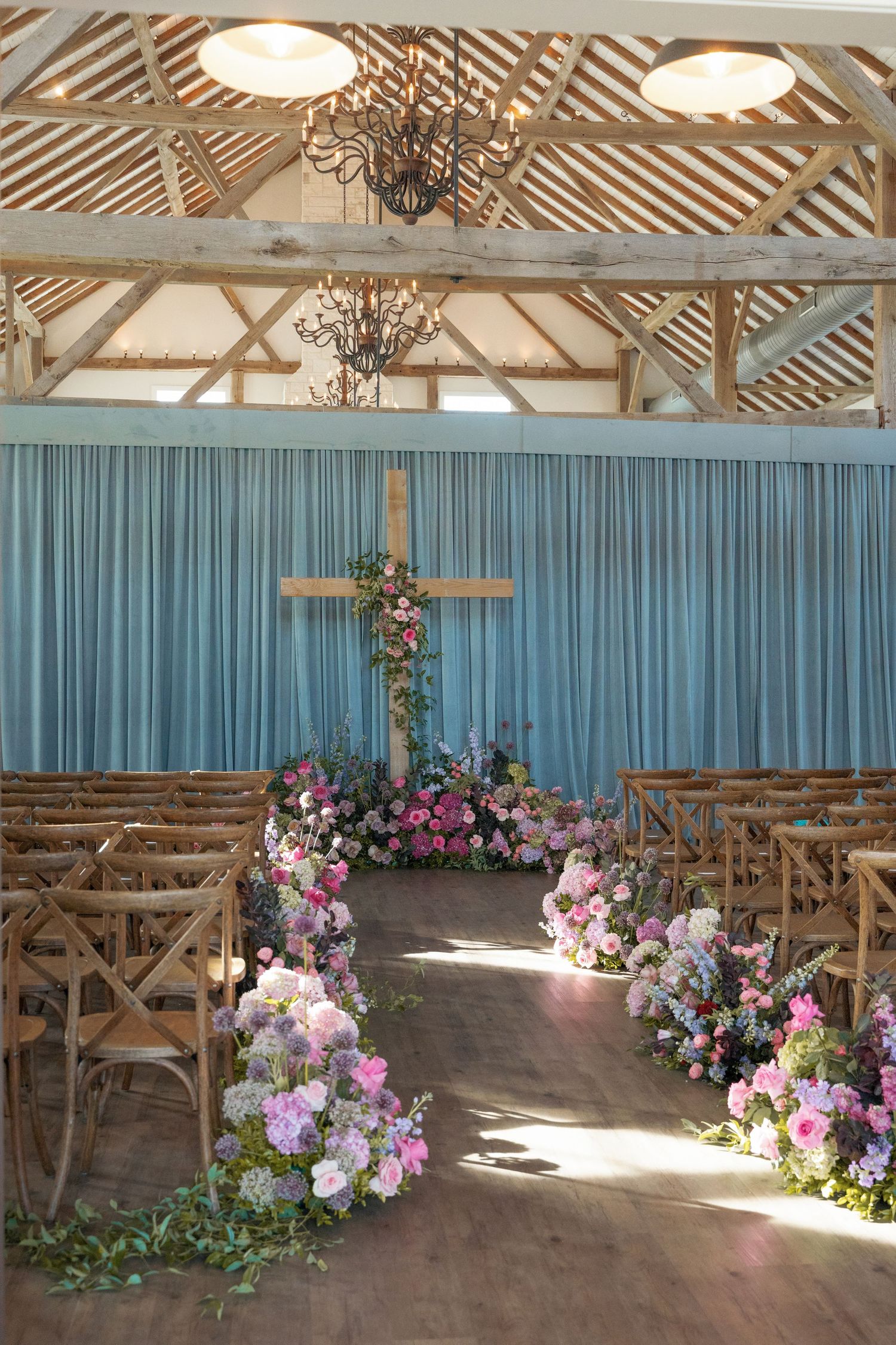 Rustic barn wedding ceremony setup with wooden chairs, blue curtain backdrop, and pink and purple floral arrangements along aisle.