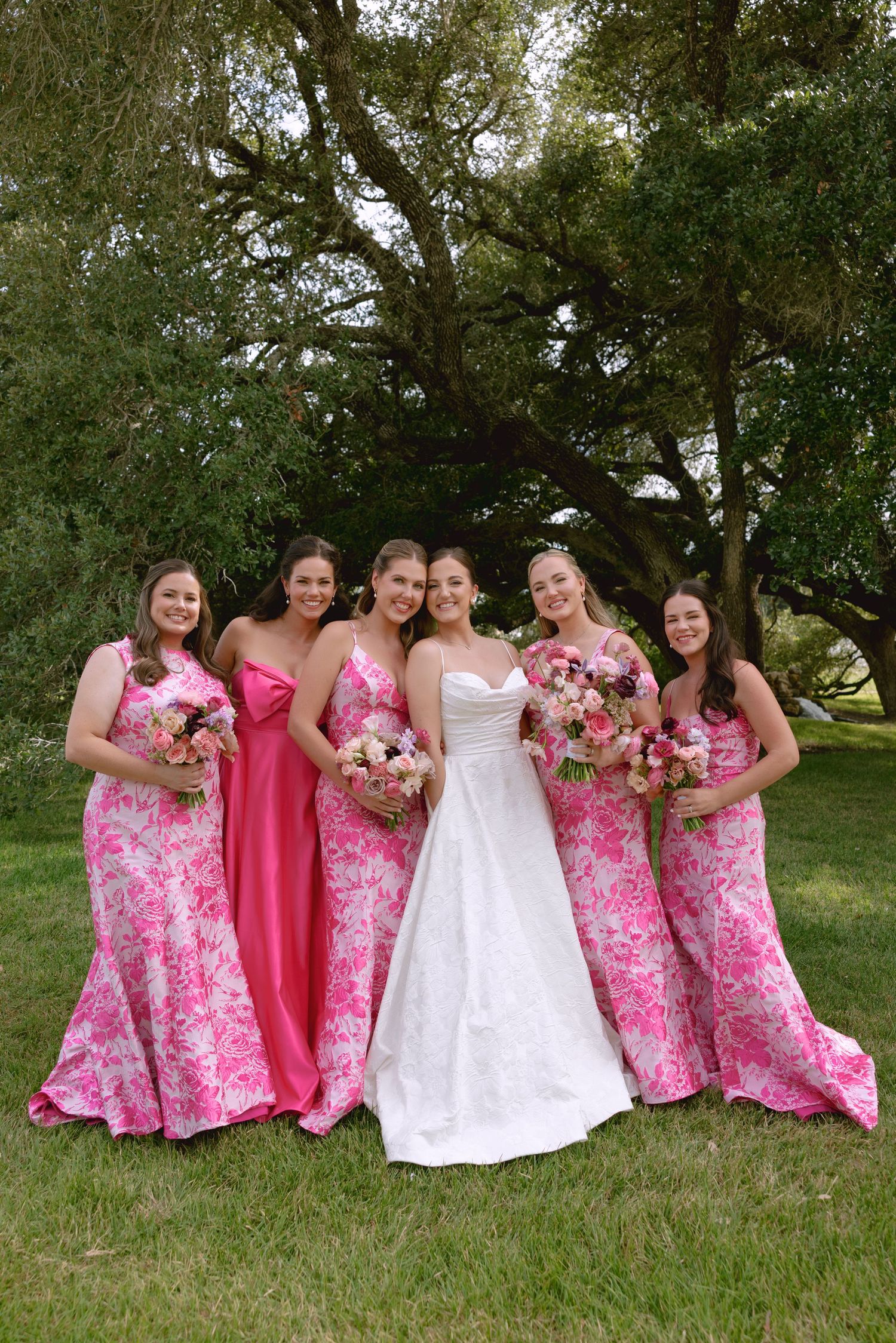 Bridesmaids in pink floral and solid dresses pose playfully on grass with the bride in white.