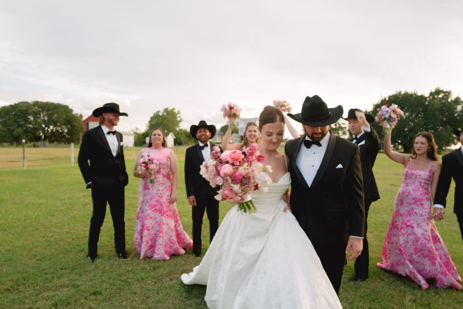 Wedding party in pink bridesmaid dresses and black tuxedos pose playfully on a green lawn for photos.