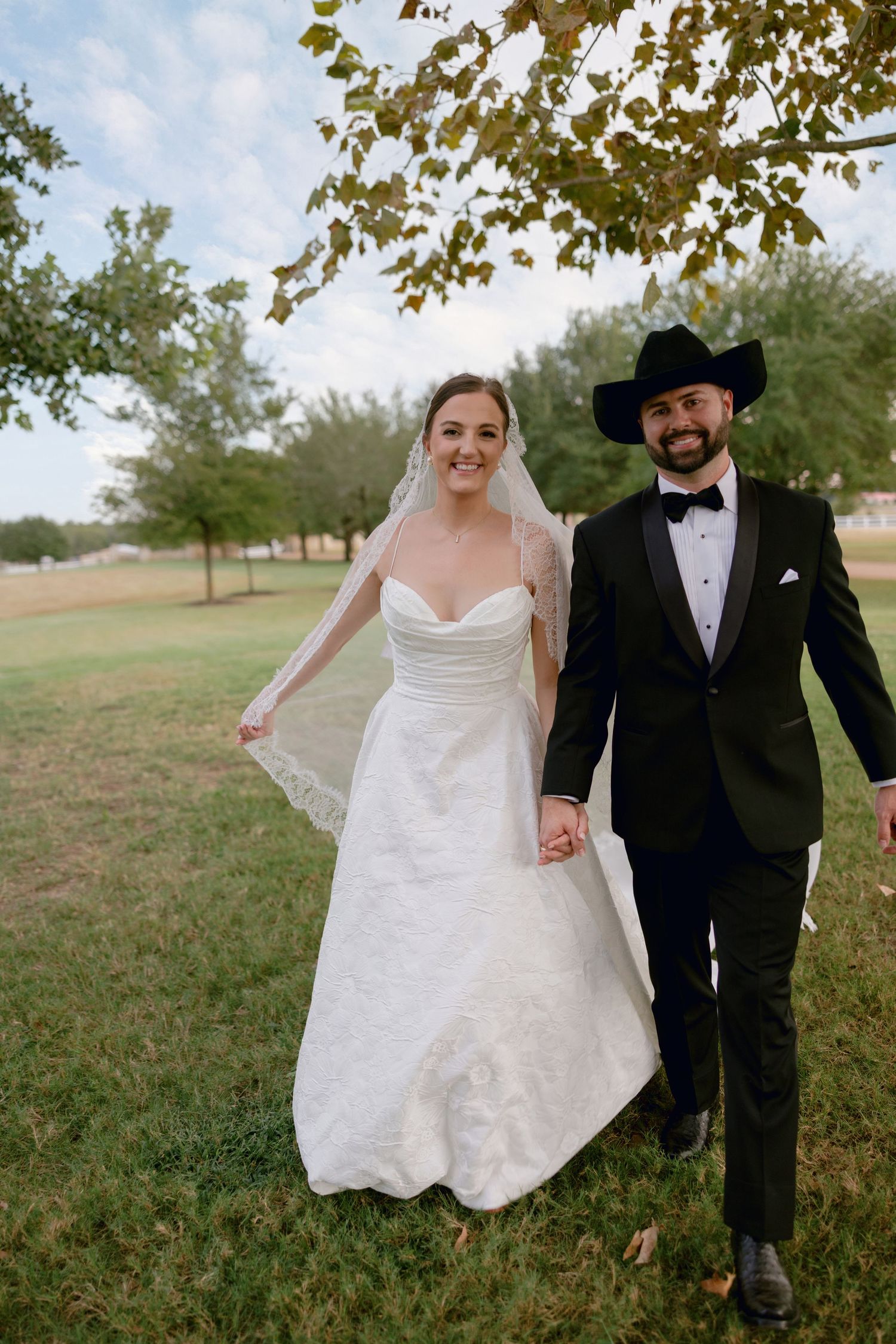 Couple walks and poses together outdoors, with bride in white dress and groom in black tuxedo.