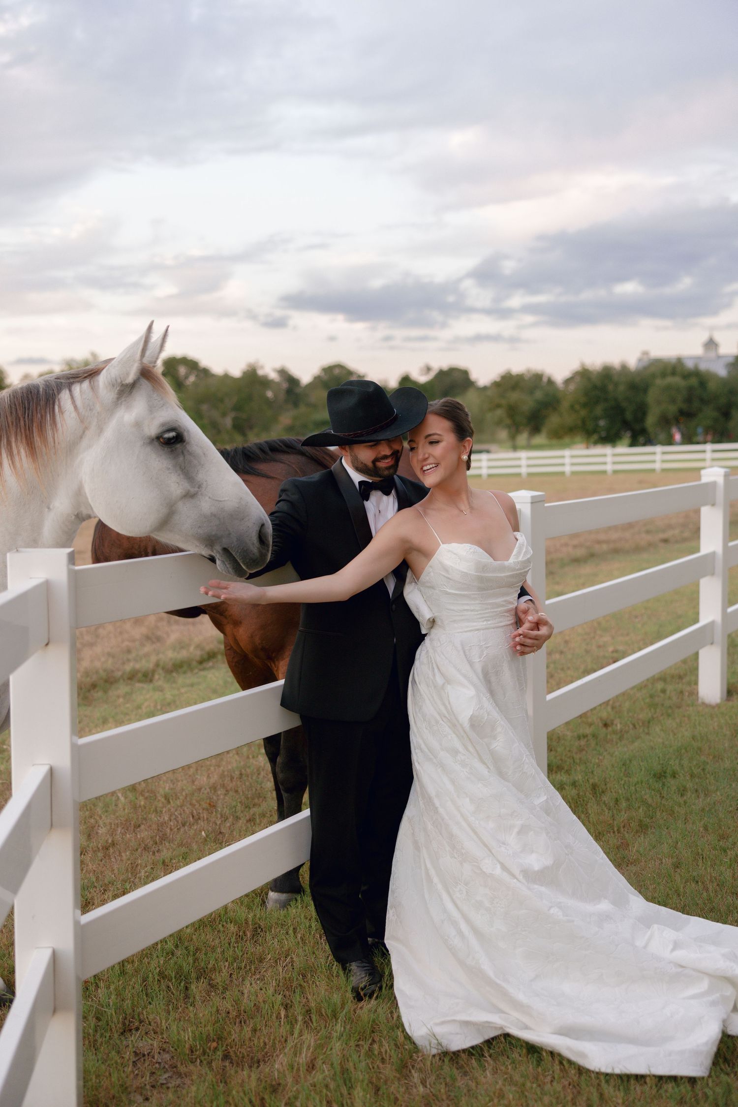 A couple in wedding attire poses for romantic photos with horses along a white fence at sunset.