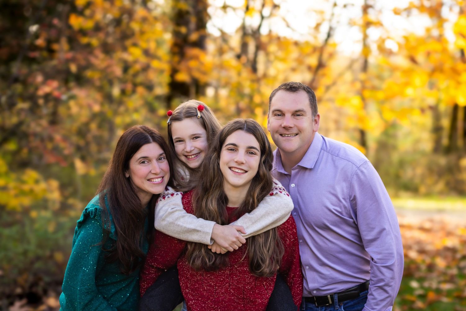 A group of four people pose together outdoors during autumn with golden fall foliage in the background.