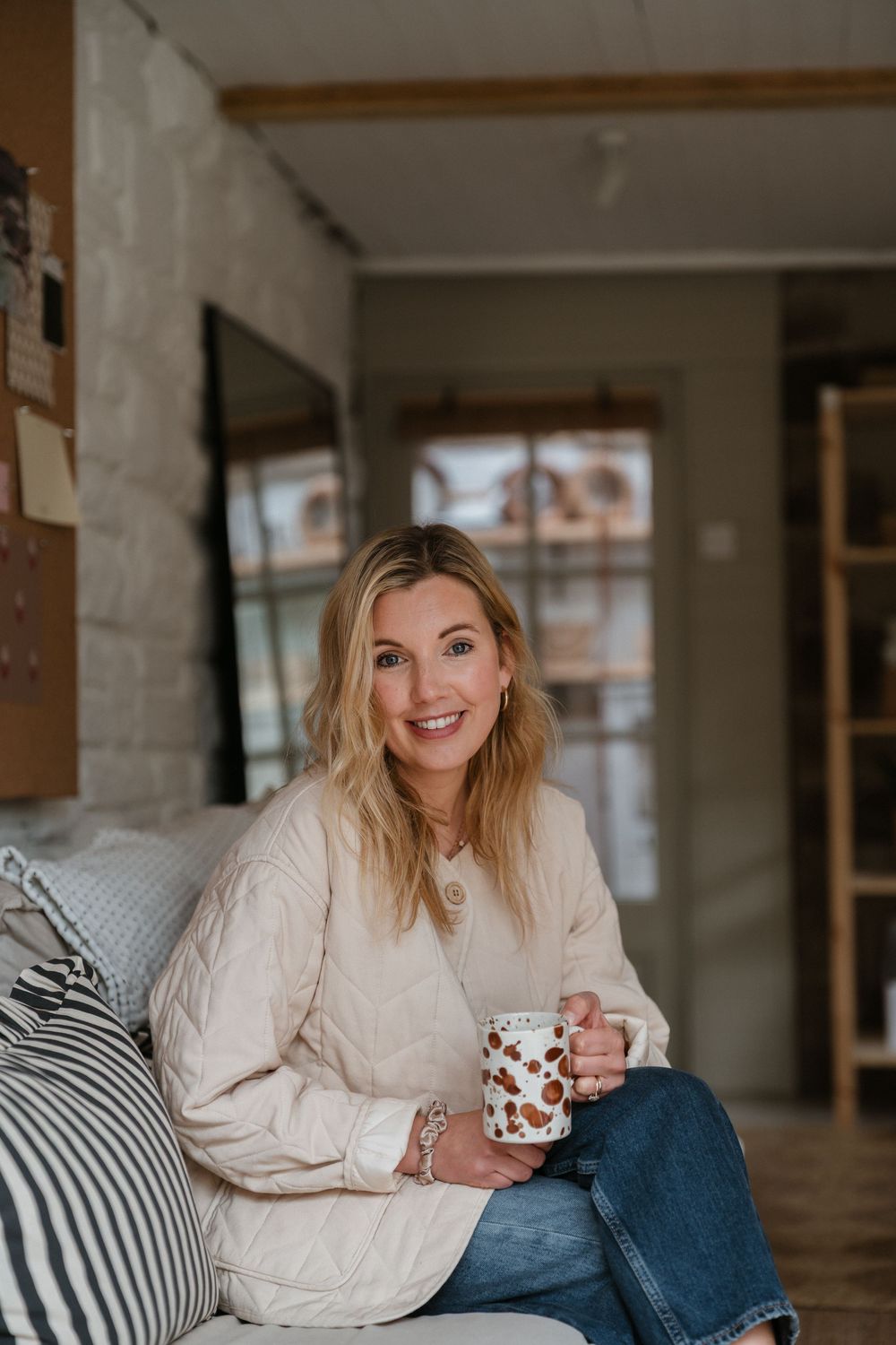 Person relaxing on couch wearing cable knit sweater and jeans while holding coffee mug in modern rustic room.