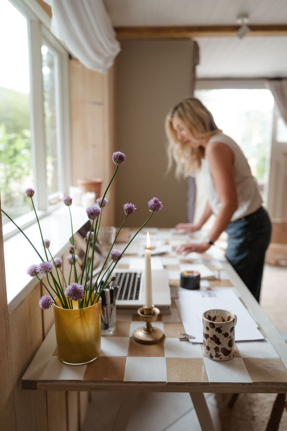 Purple chive flowers in a yellow vase sit on a wooden table while someone works in the background at home.