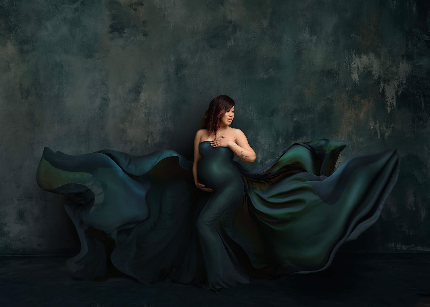 Artistic fashion portrait against dark textured wall with flowing emerald green fabric creating dramatic movement.