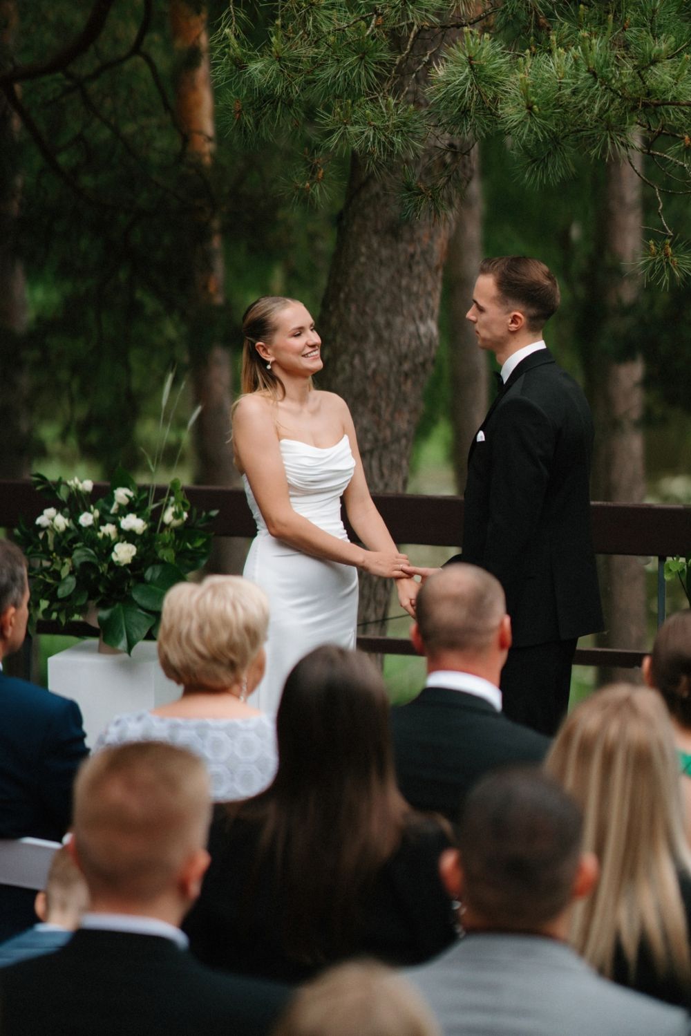 Outdoor wedding ceremony in a pine forest with guests seated on white chairs.