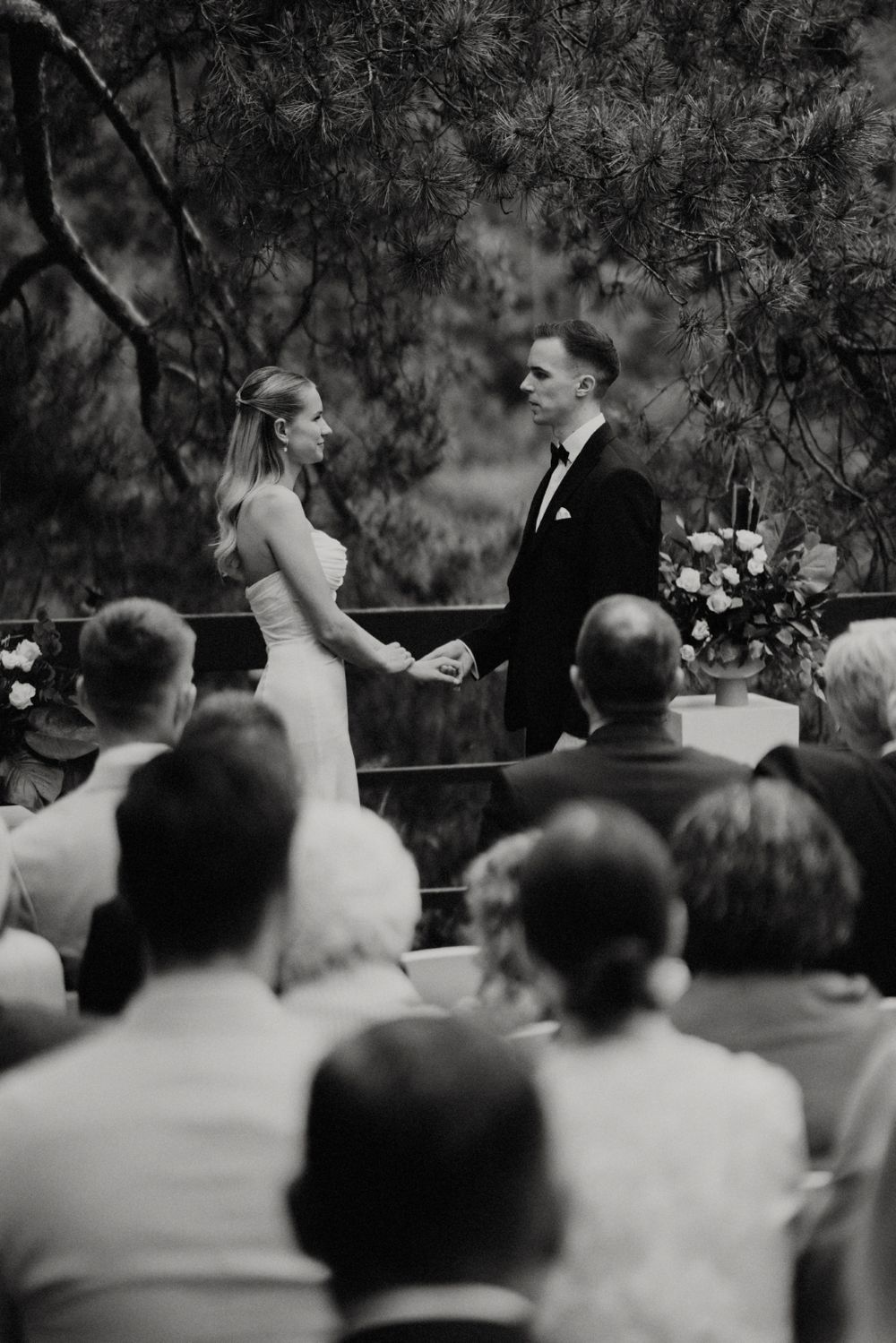 Black and white photo of an outdoor wedding ceremony with pine trees in the background.