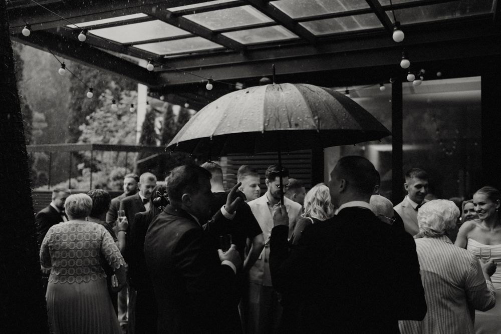 Black and white photo of wedding guests gathered under a covered patio with umbrellas.