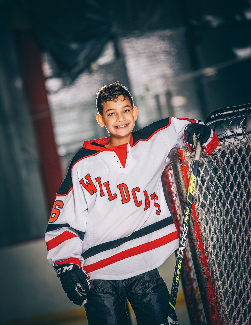 Young hockey player in a white and red Wildcats jersey smiles while leaning against the rink boards.