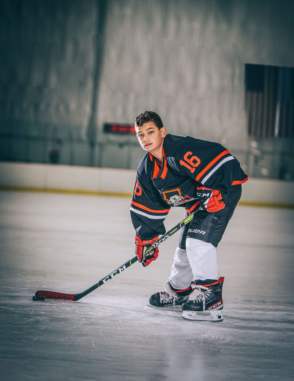 Hockey player in black and orange jersey number 16 crouches on ice rink with stick in ready position.
