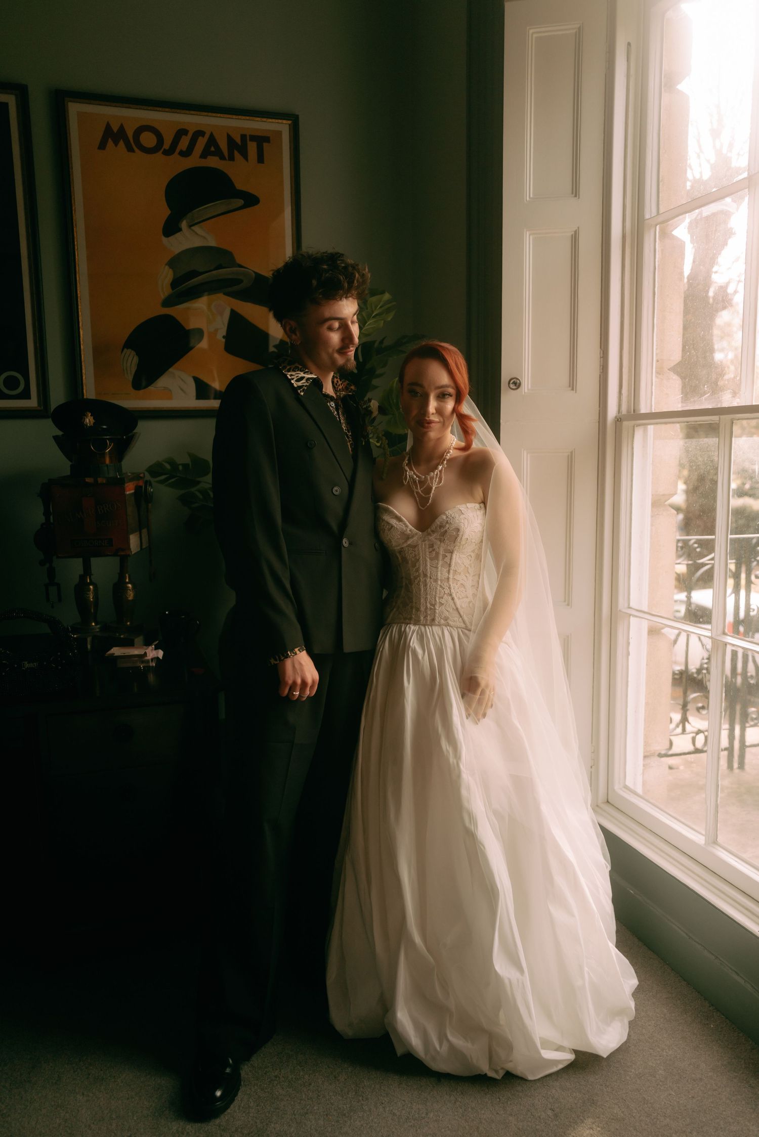 A couple in wedding attire share an intimate moment near a window in moody lighting.