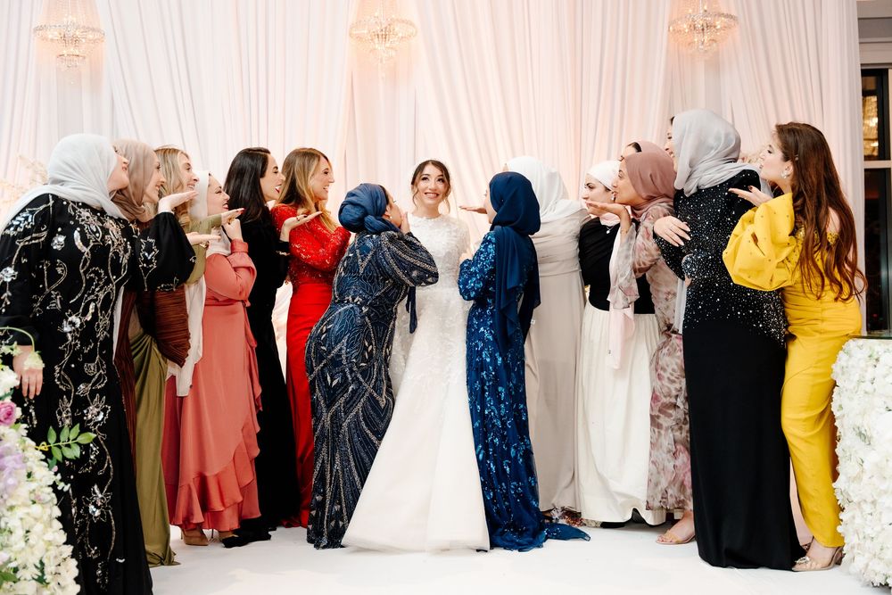 Muslim bride in white gown celebrates with group of female guests in colorful hijabs at wedding.