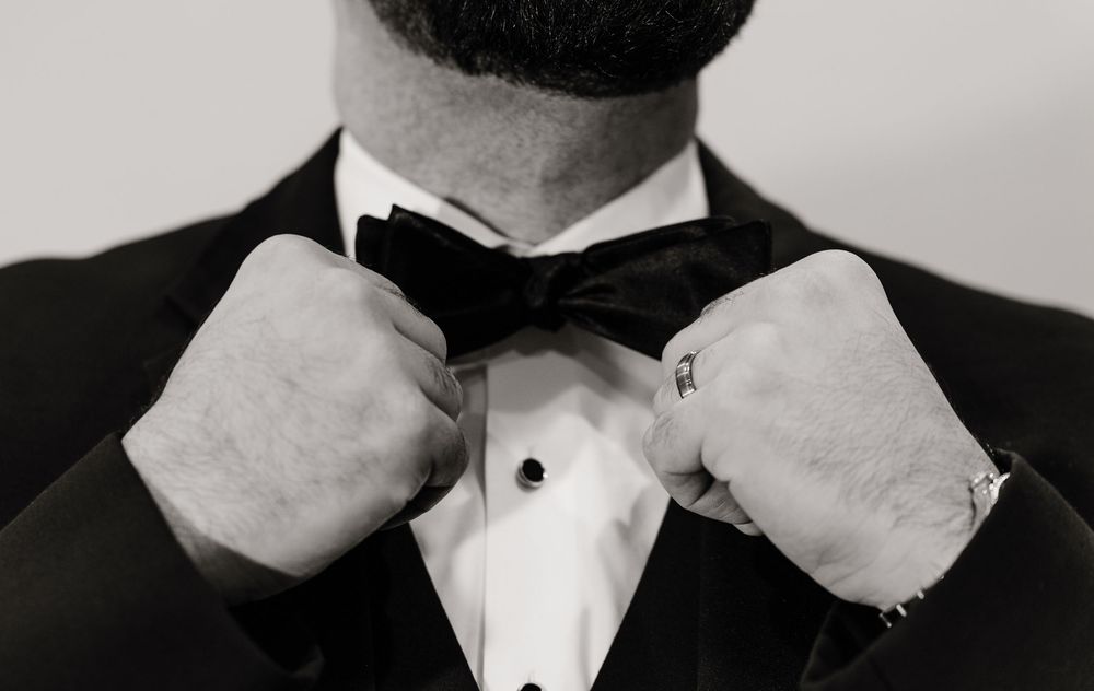 Groom in black tuxedo adjusts bow tie in black and white wedding portrait photo.