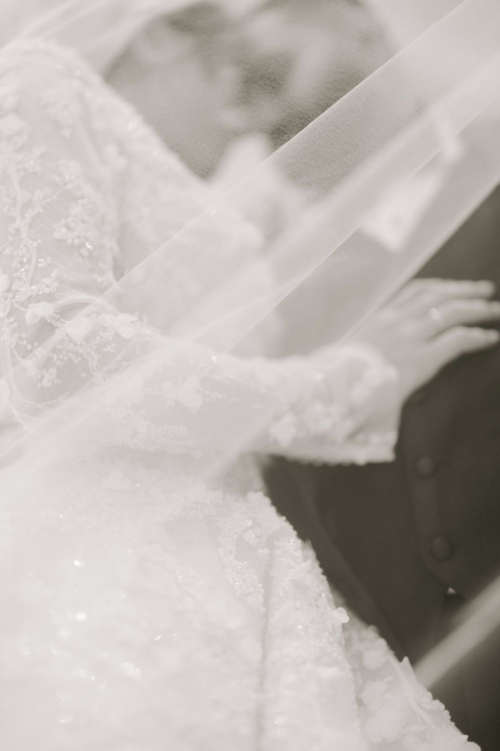 Close-up of brides white beaded lace wedding dress and flowing veil in soft monochrome photo.