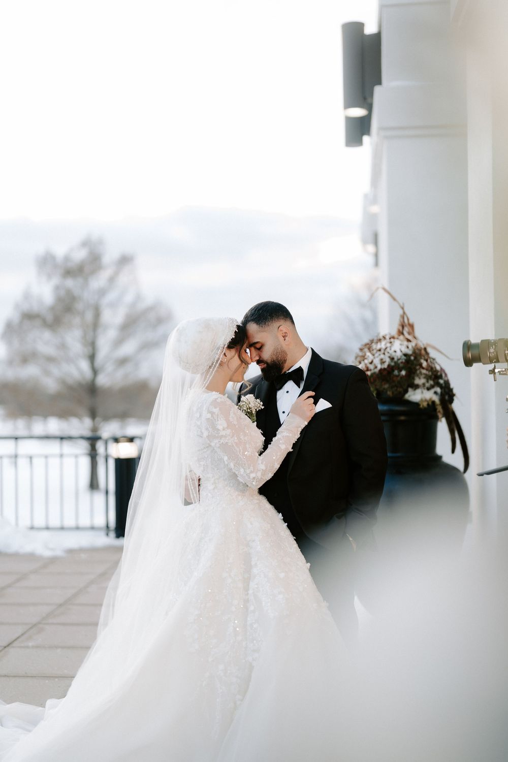 Bride in white lace gown and groom in black tuxedo share romantic moment in snowy winter wedding.