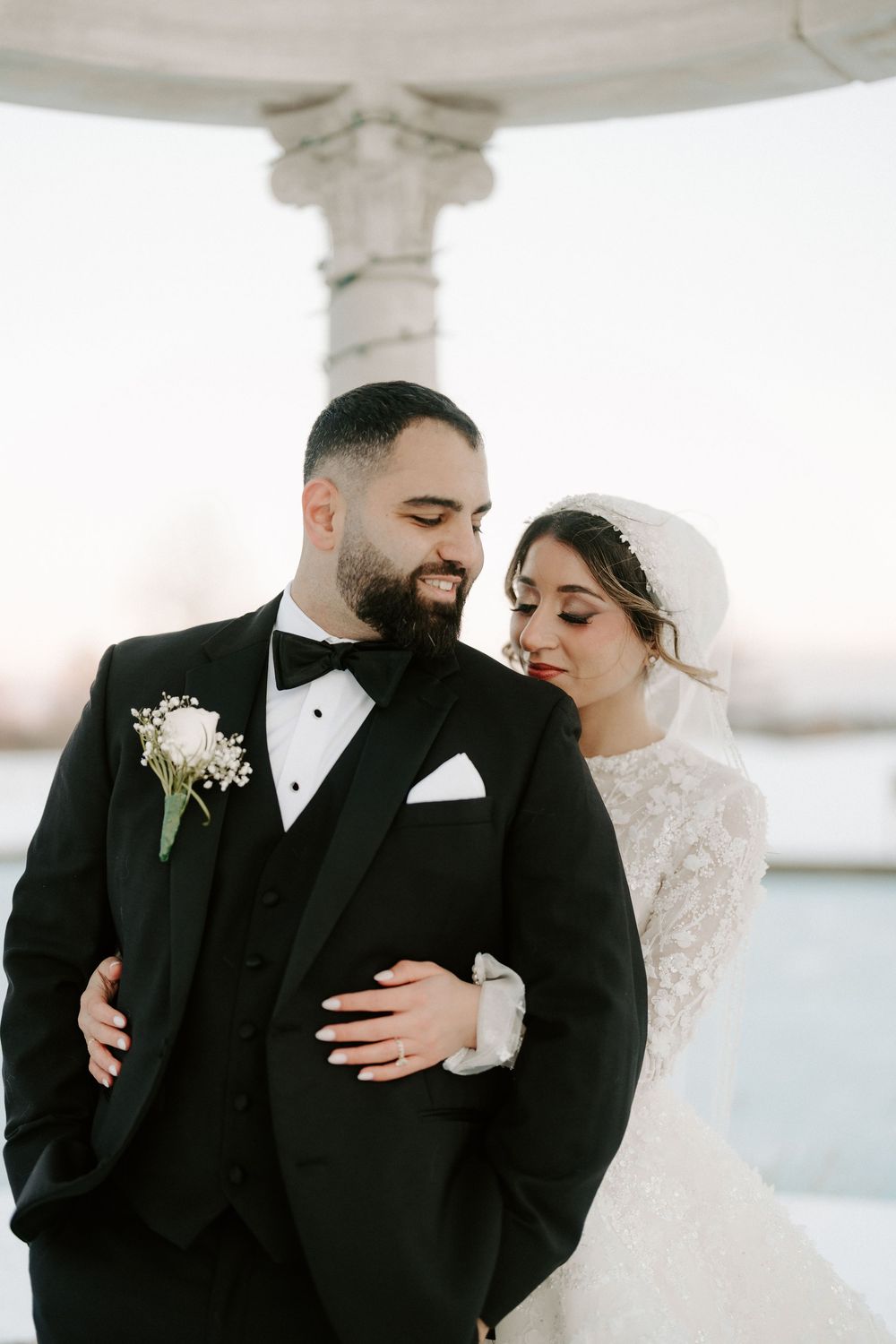 Bride and groom sharing an intimate moment under a white gazebo, groom in black tuxedo with boutonniere, bride in lace wedding gown.