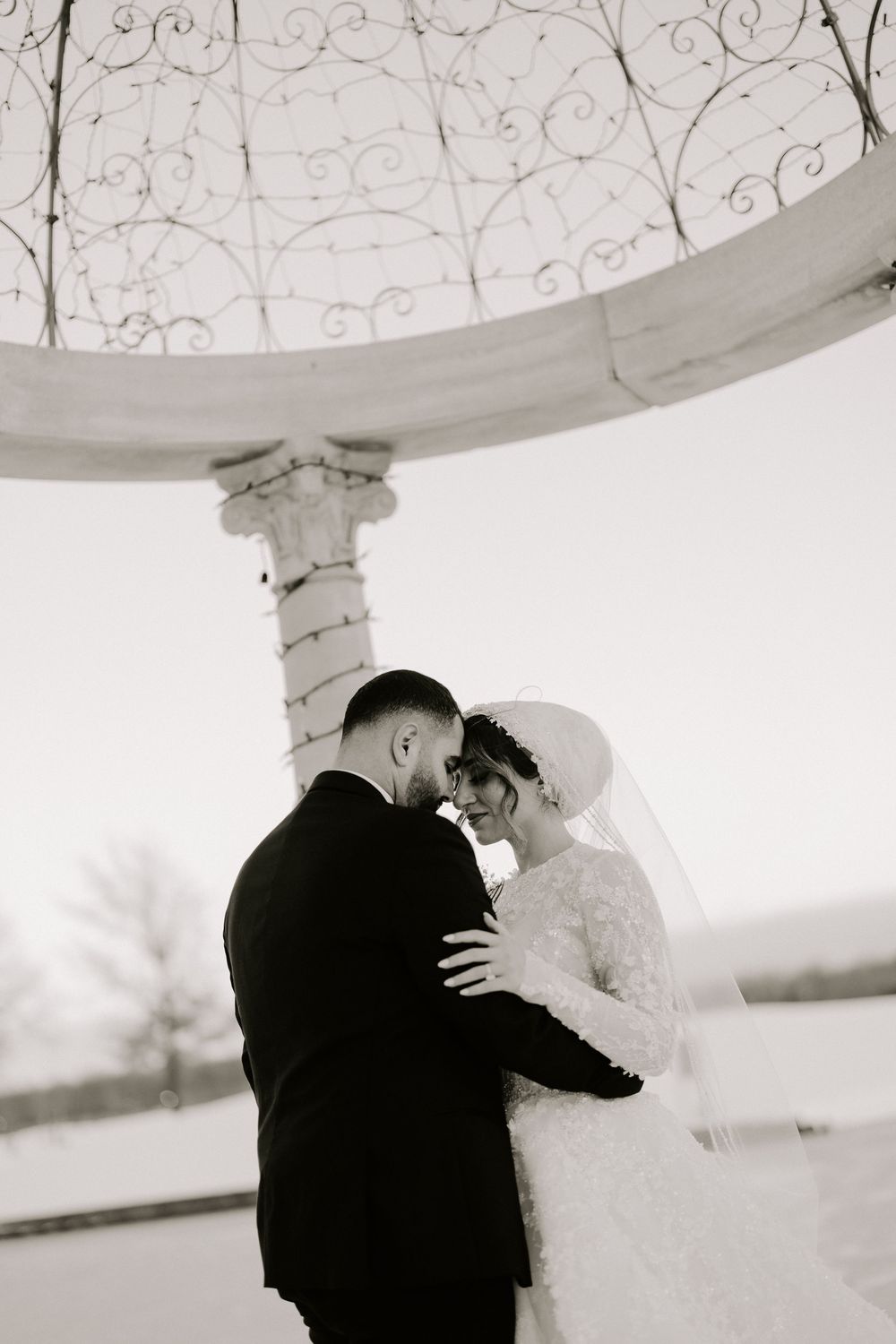 Black and white photo of bride and groom embracing under ornate iron gazebo, touching foreheads romantically in winter setting.