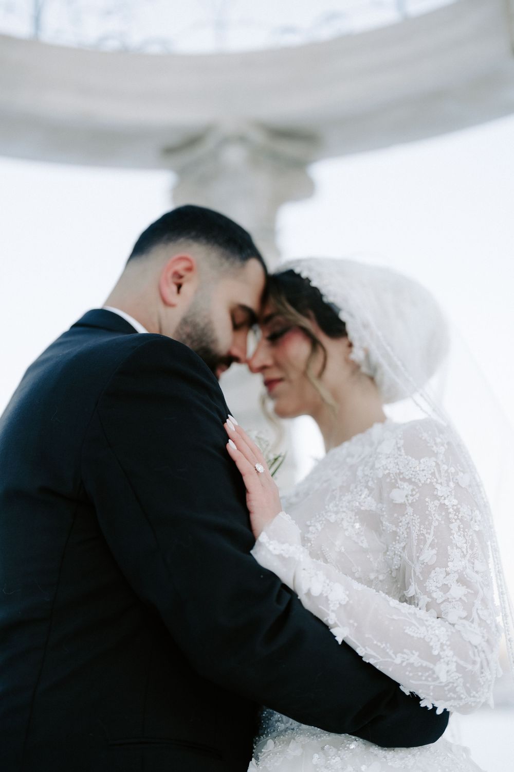 Close-up black and white photo of bride and groom nearly kissing, bride in lace long-sleeve gown, groom in black suit.