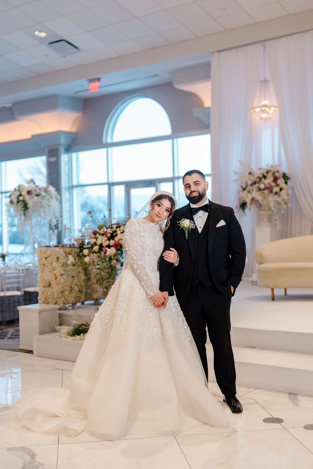 Bride and groom posing in elegant ballroom with floral arrangements, bride in ball gown, groom in black tuxedo holding hands.