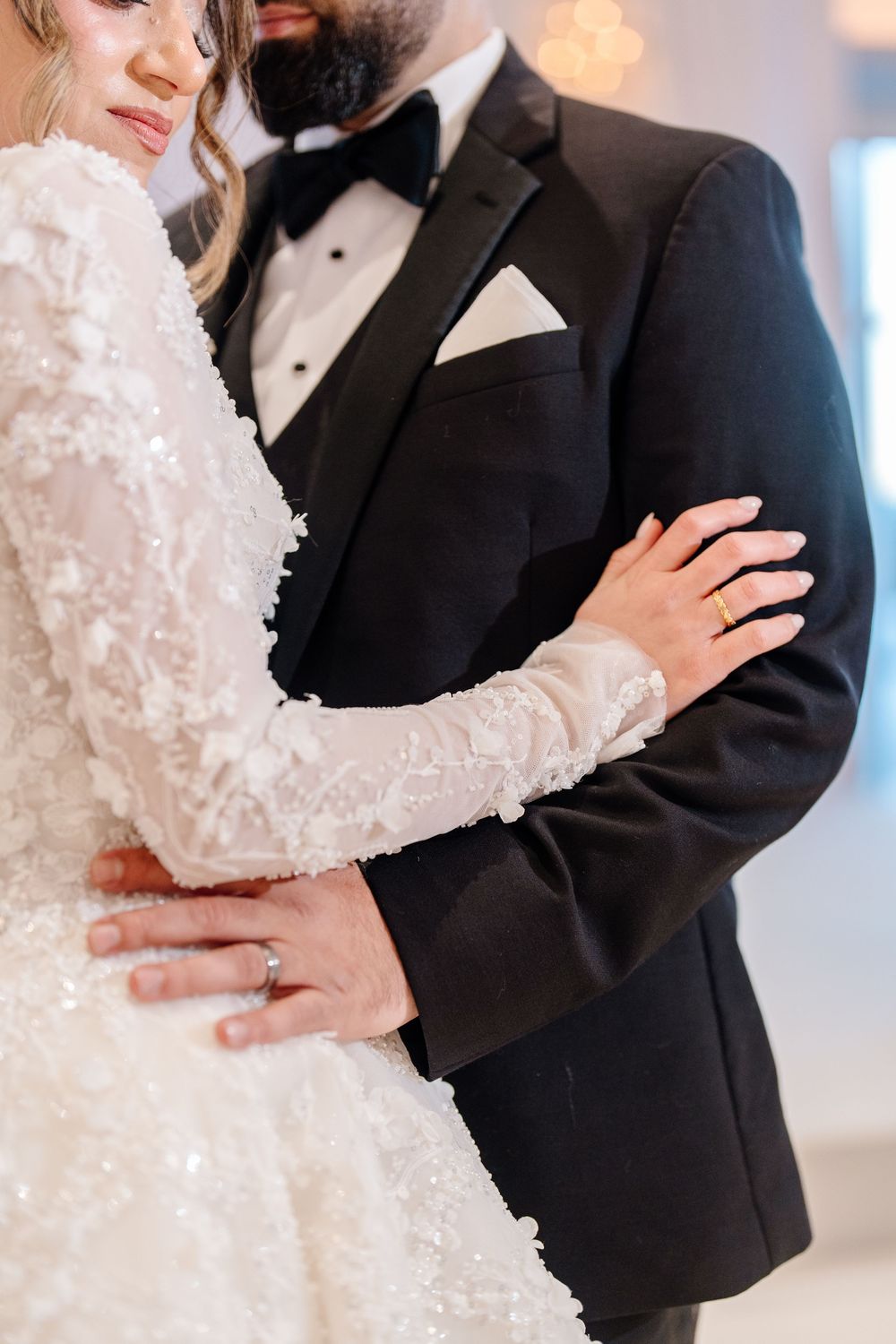Bride in beaded lace long-sleeve gown embraces groom in black tuxedo at wedding ceremony.