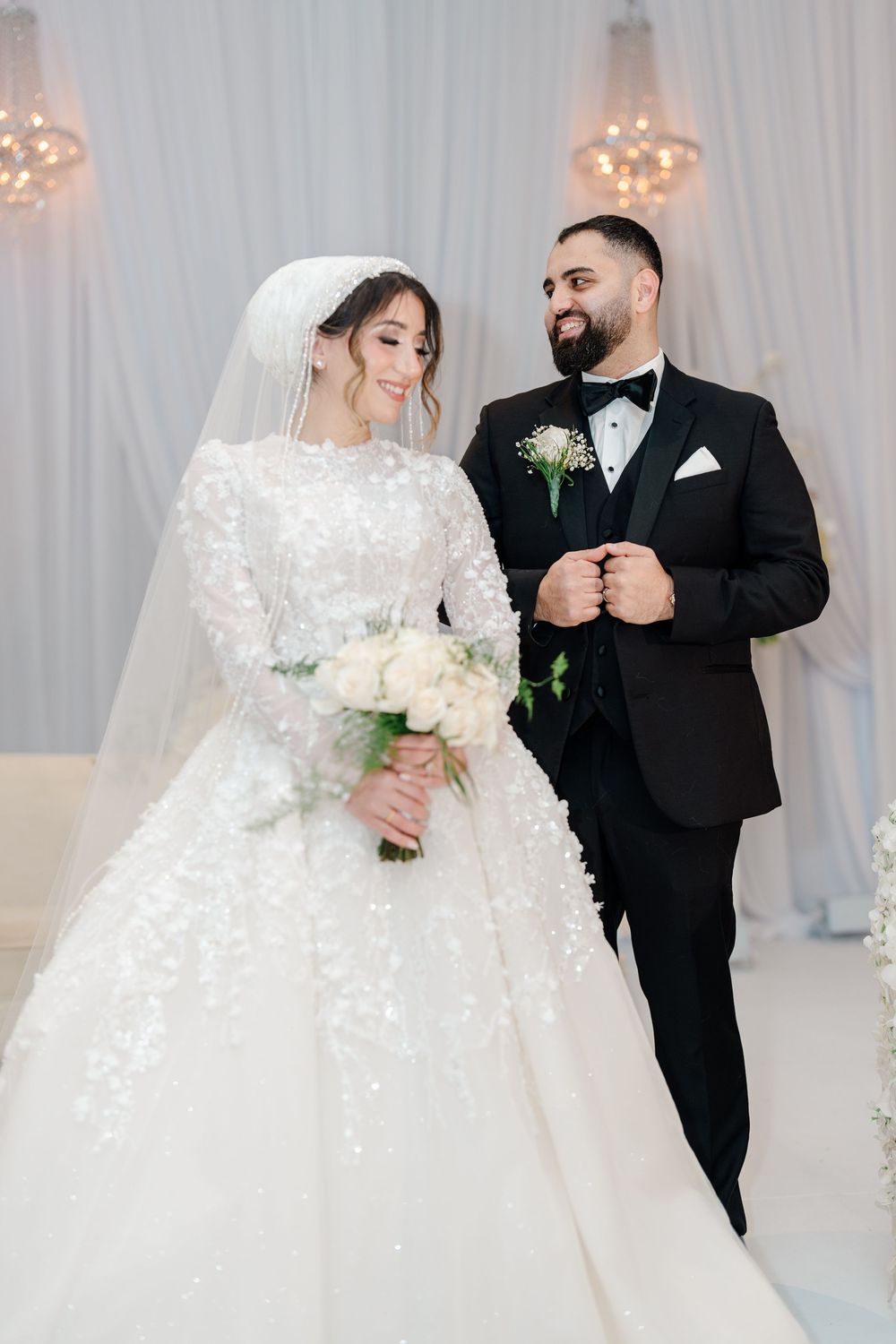 Bride in white beaded ball gown with veil holds bouquet beside smiling groom in black tuxedo.