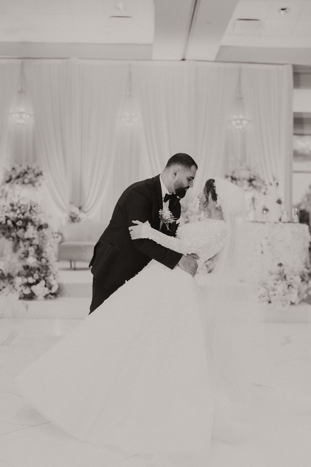 Black and white photo of groom dipping bride during first dance in decorated ballroom with white draping and floral arrangements.