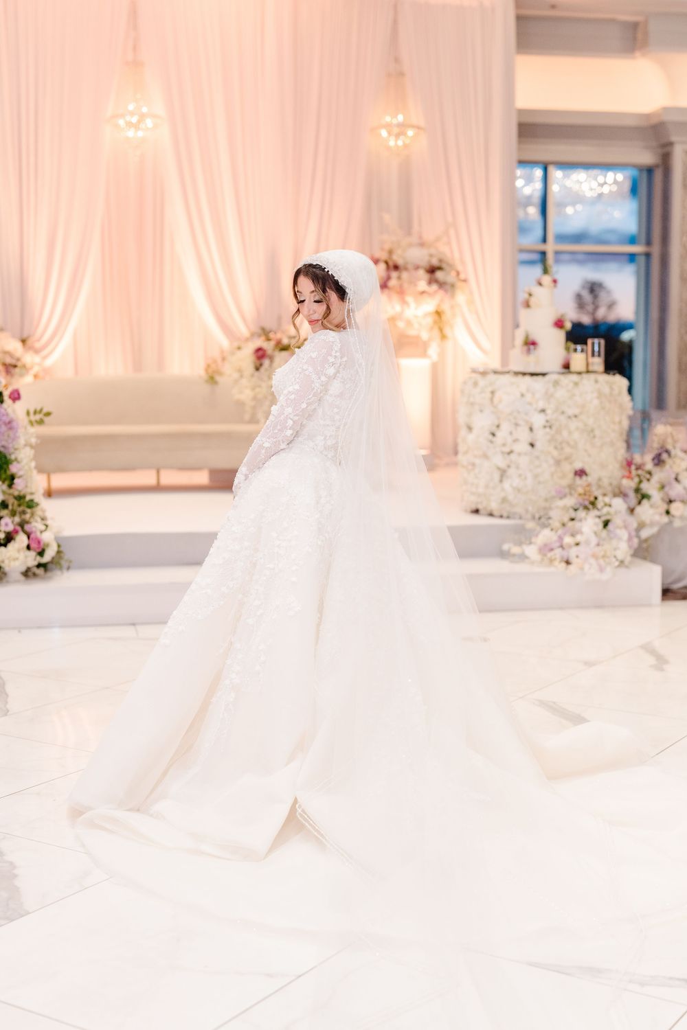 Bride in elegant lace ball gown with cathedral veil twirling on white dance floor in beautifully decorated white wedding reception venue.