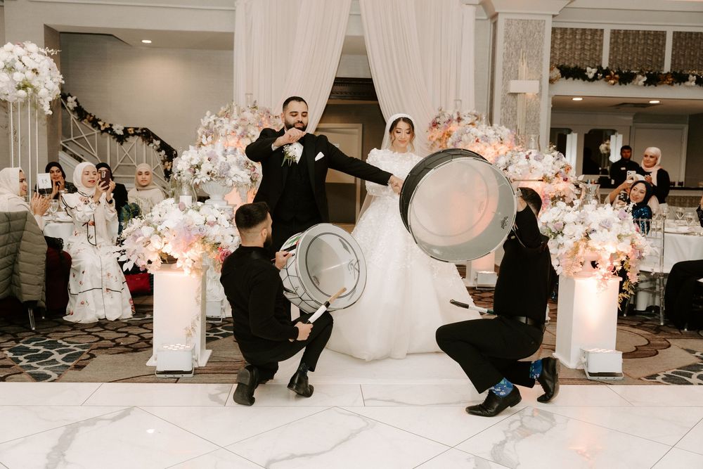 Bride and groom celebrate at wedding reception with drummers performing on marble dance floor amid white floral decor.
