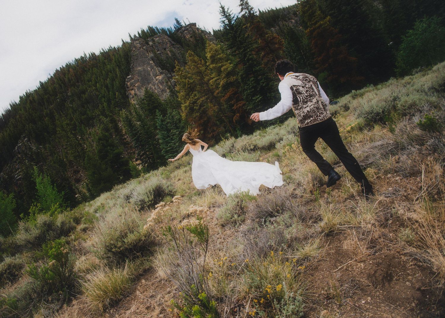 A couple walks through a vibrant field of yellow wildflowers with mountains in the background.