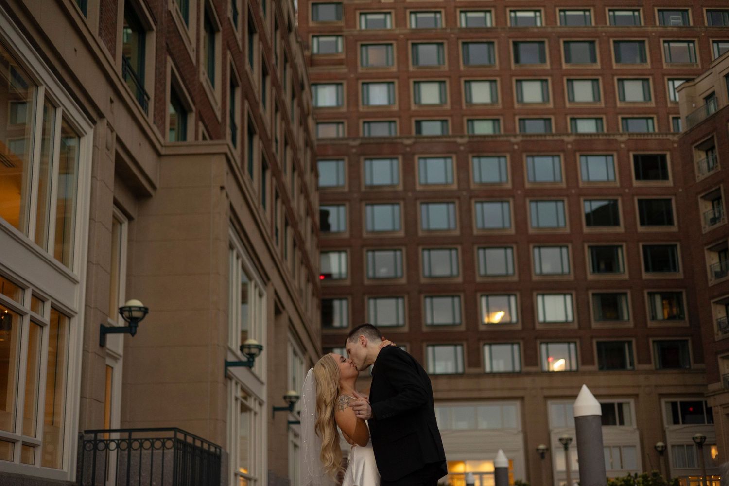 A romantic evening moment between two people outside a tall brick building with illuminated windows in the background.