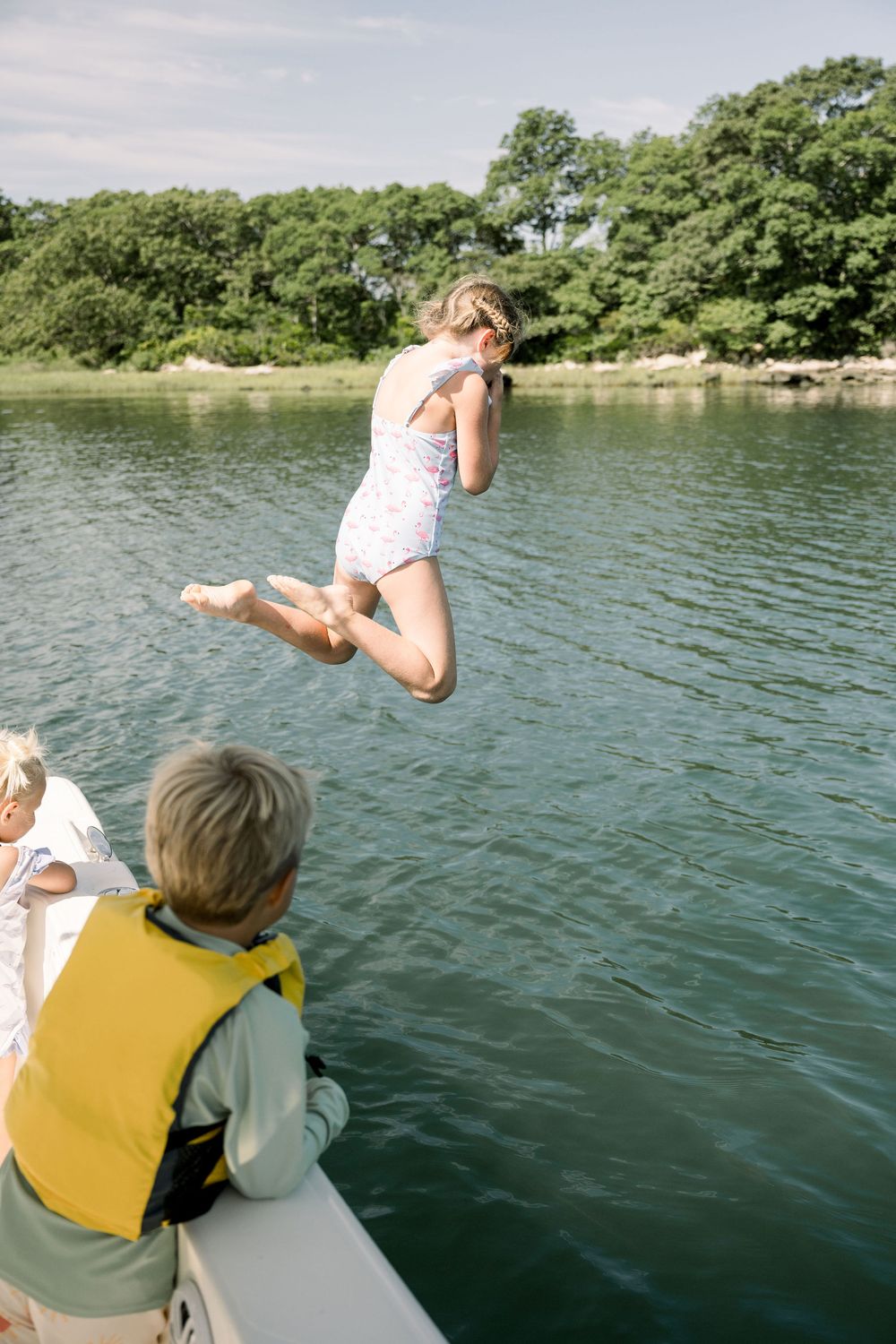 Someone jumps off a boat into a calm lake surrounded by green trees on a summer day.