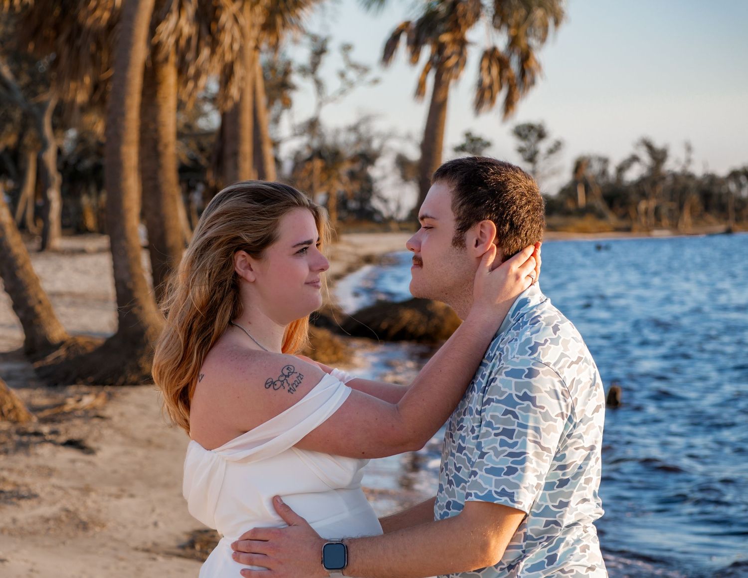 A couple embraces by a waterfront at sunset with palm trees lining the shore.