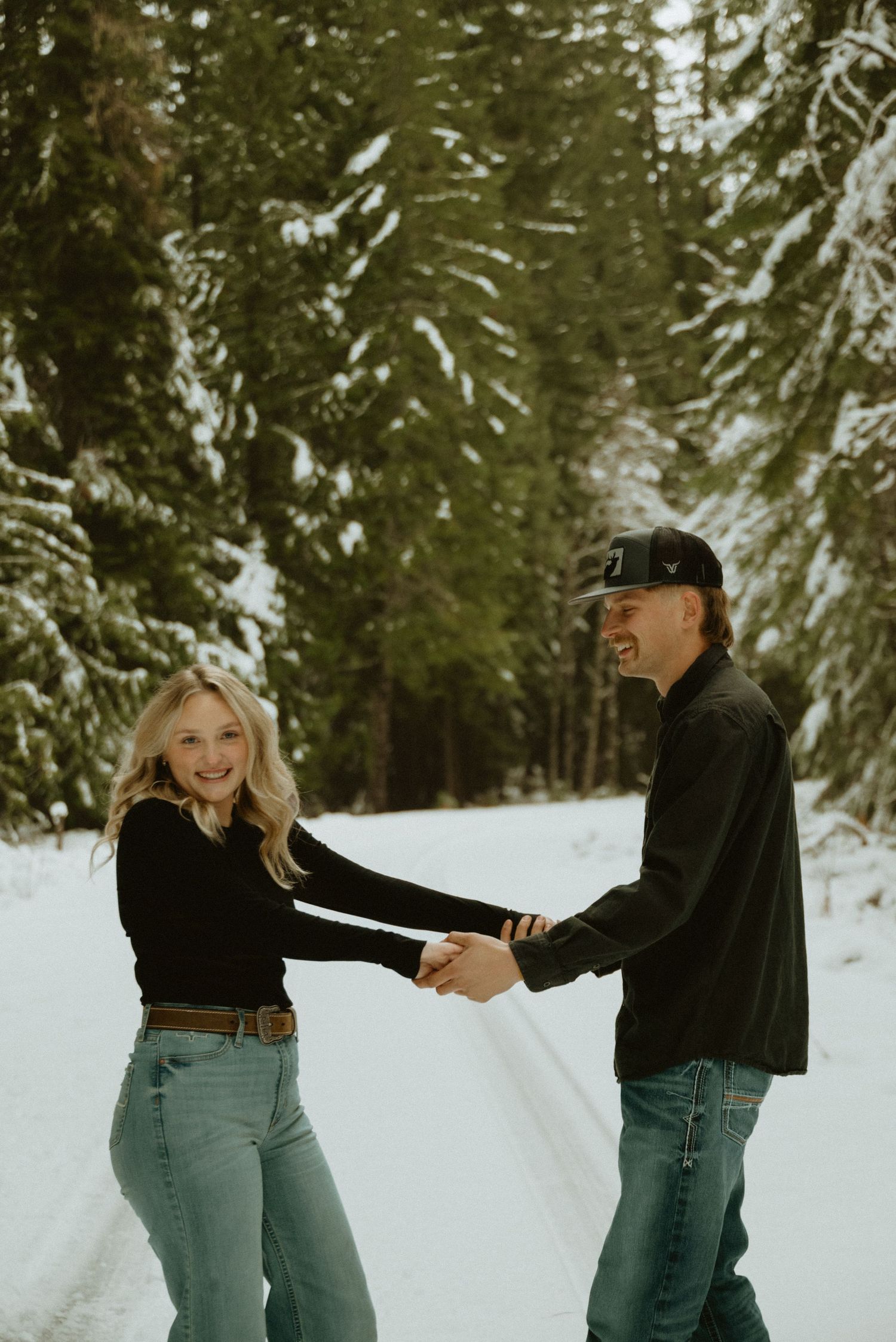 A couple in winter attire playfully dances together on a snow-covered path surrounded by evergreen trees.