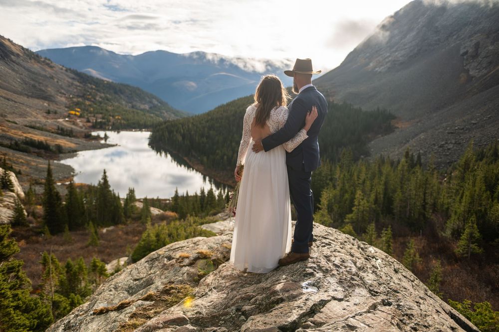 An eloping couple stand in an embrace looking out over the overlook at an alpine lake surrounded by mountains and bright sunrise light.