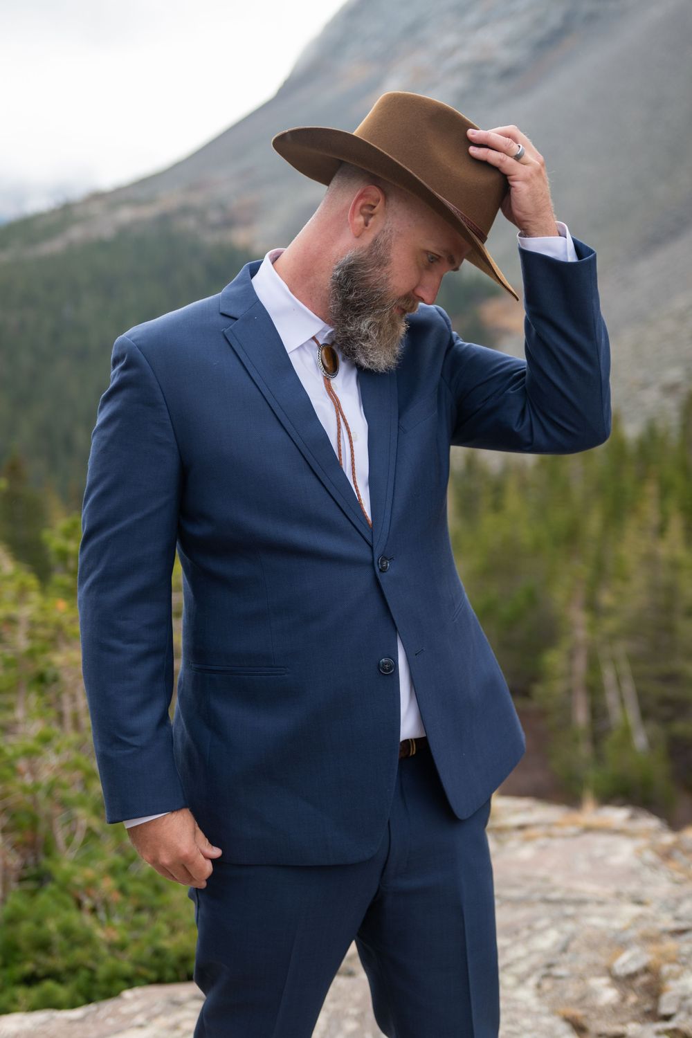 A groom poses for his wedding shot holding his cowboy hat in formal attire. He has mountains and pine trees in the background.