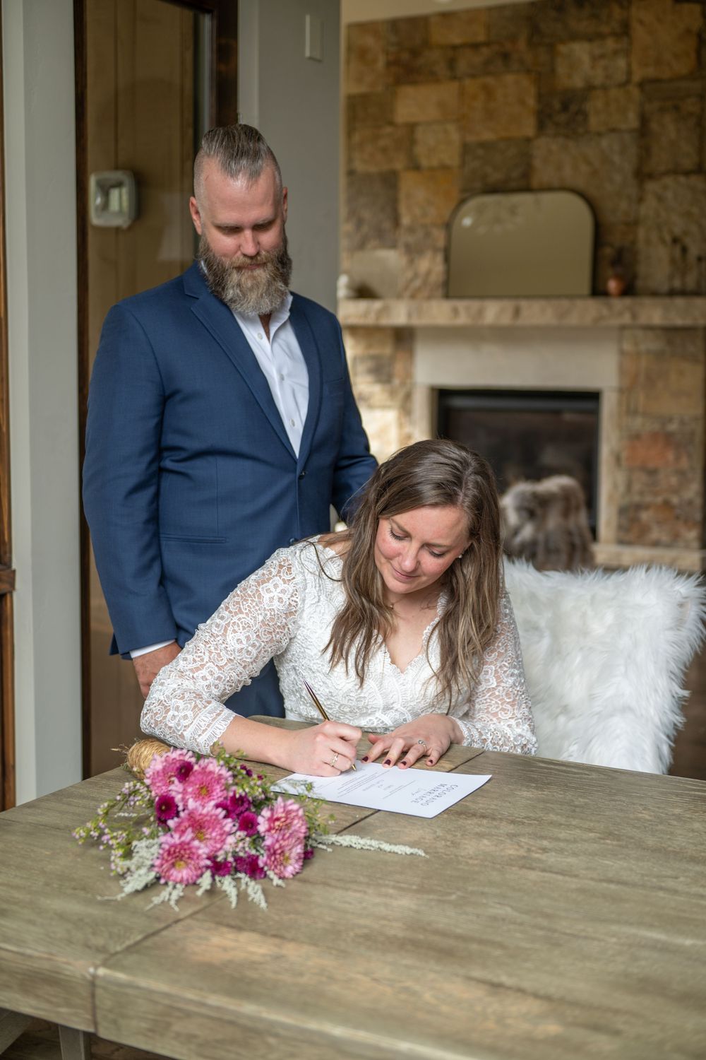 A bride and groom sign their marriage license in their vacation rental. Her bouquet is on the table in front of her. He stands behind her.