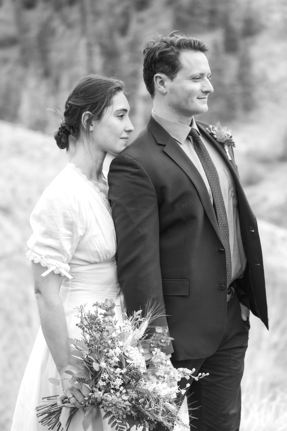 A black and white photo showing an eloping couple in formal attire with the bride holding a rustic wildflower bouquet. She stands behind him