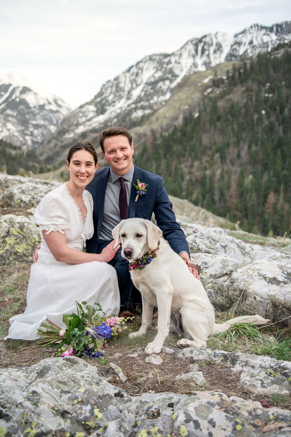 An eloping couple poses with their dog on rocky mountain terrain with the snow-capped peaks of the San Juan Mountains in the background.