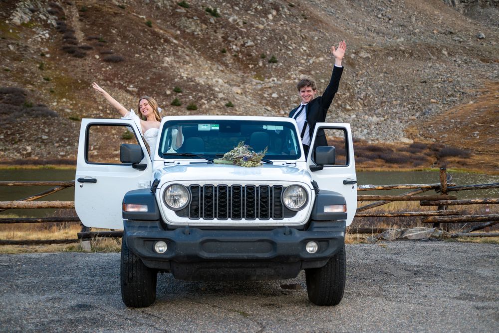 A bride and groom celebrate with their arms in the air out of the open top of a Jeep. They are surrounded by mountain scenery .