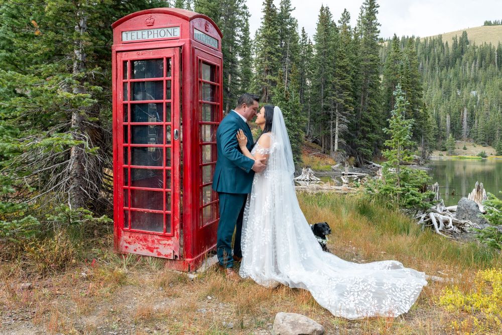 A bride and groom share a kiss near a classic red British phone booth surrounded by mountain wilderness and evergreen trees.