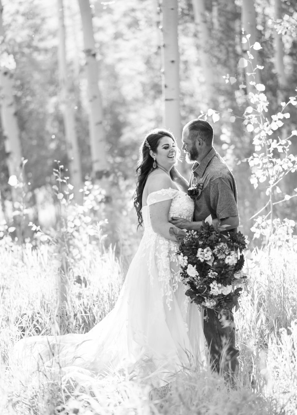 A black and white image featuring a bride and groom smiling and embracing in an brightly lit aspen grove near Ridgway.