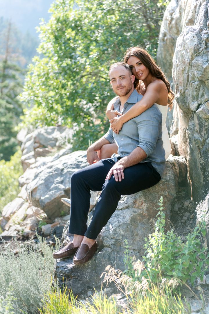 A couple embraces while sitting on rocky terrain surrounded by greenery and natural light in the rugged mountains. She is seated behind him.