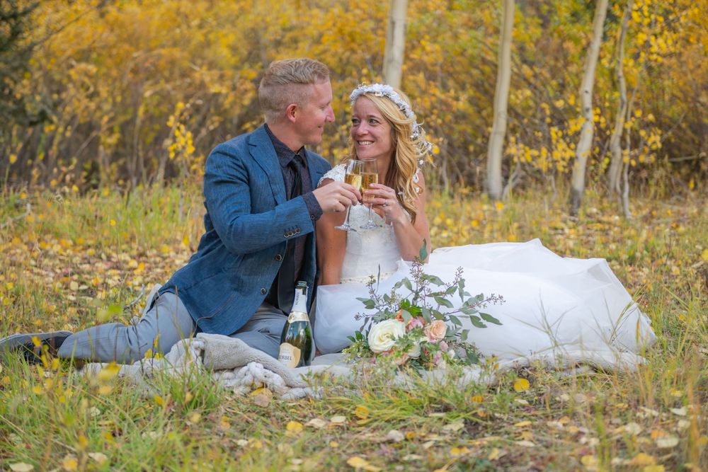 A bride and groom sit in a grassy mountain meadow together and share a champagne toast with golden aspens behind them.
