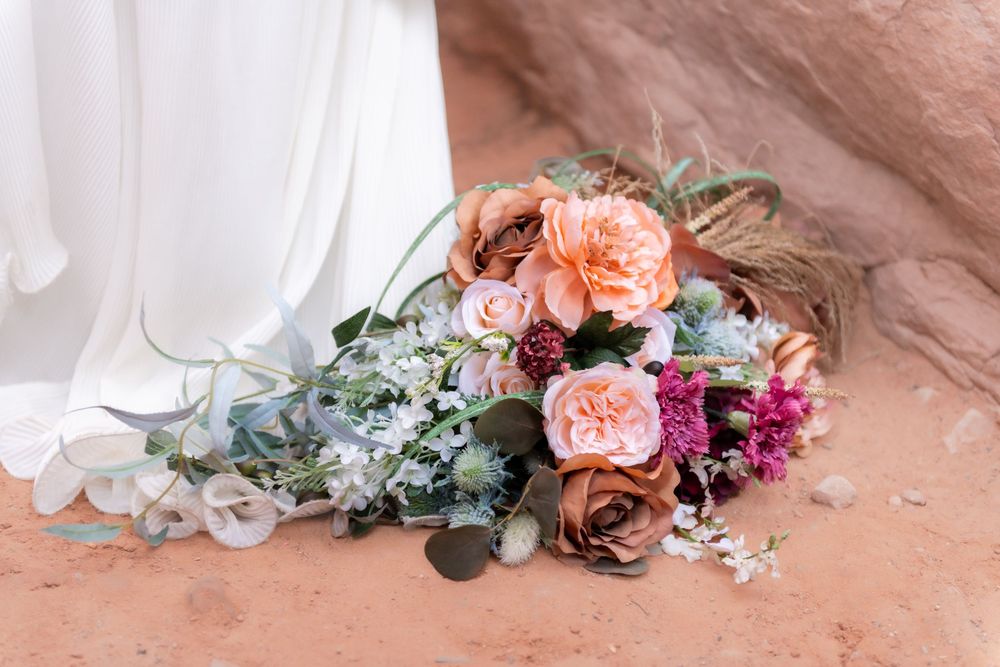 A close up shot of a bride's bouquet features soft pink and burgundy floral arrangements with eucalyptus leaves against the hem of her dress