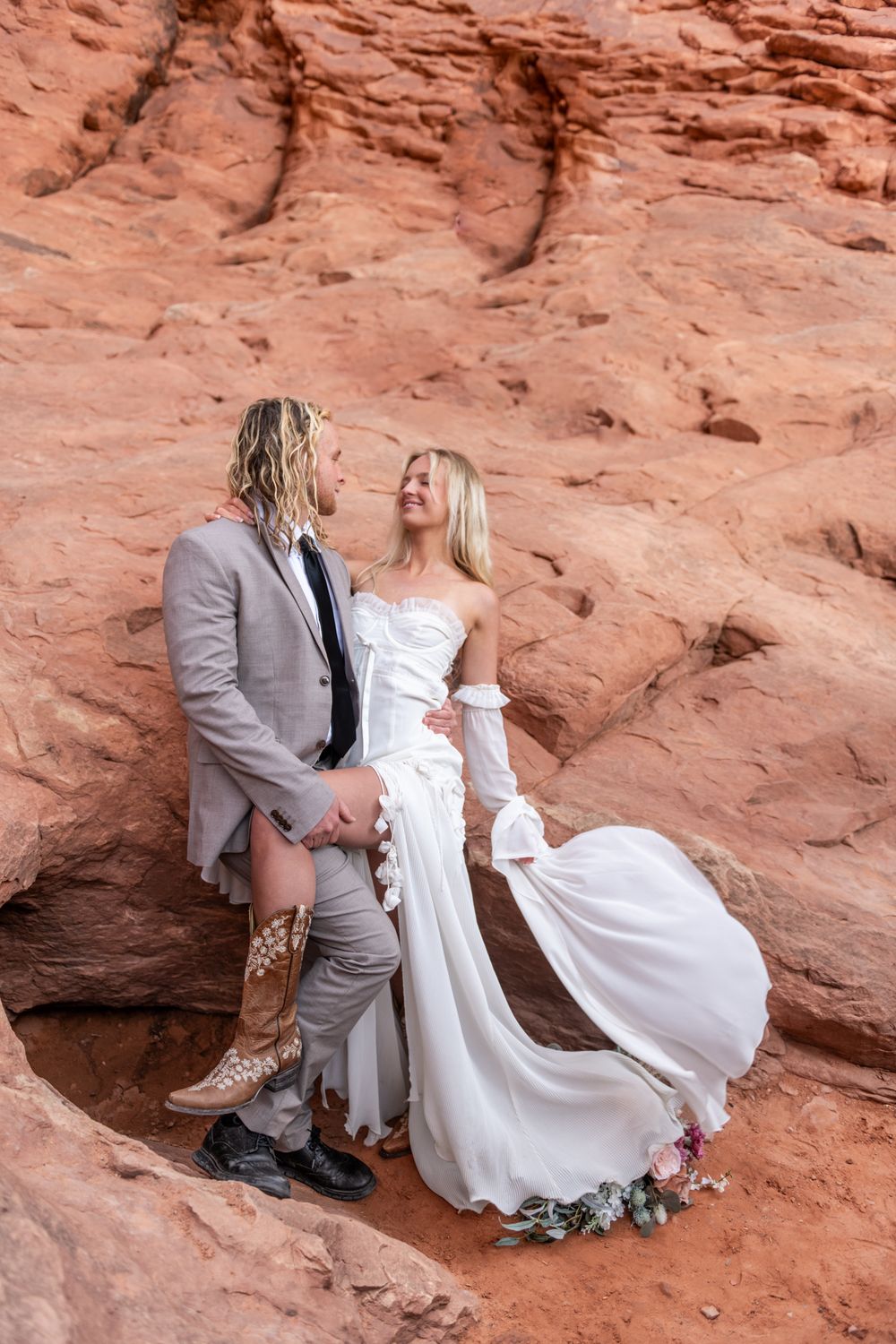 A couple in wedding attire shares an intimate embrace against dramatic red rock formations in a desert landscape.