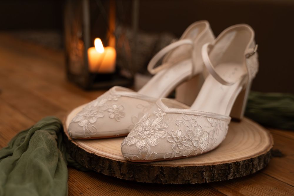 Elegant white lace bridal shoes displayed on wooden slice with candle lantern in background.
