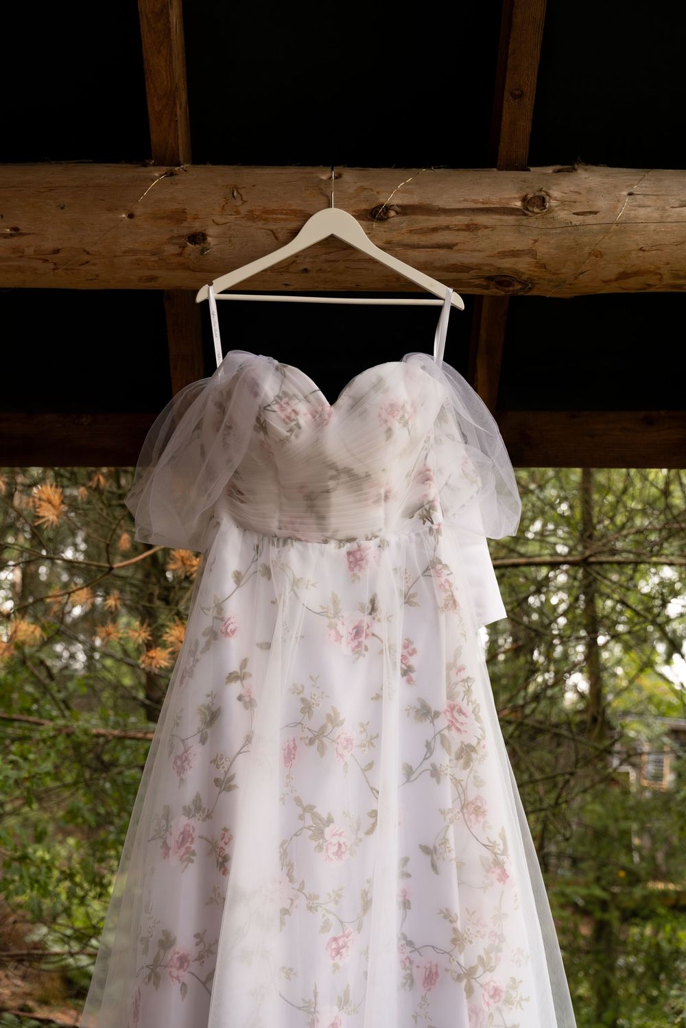 Delicate floral wedding dress hanging on white hangers against rustic wooden beams.