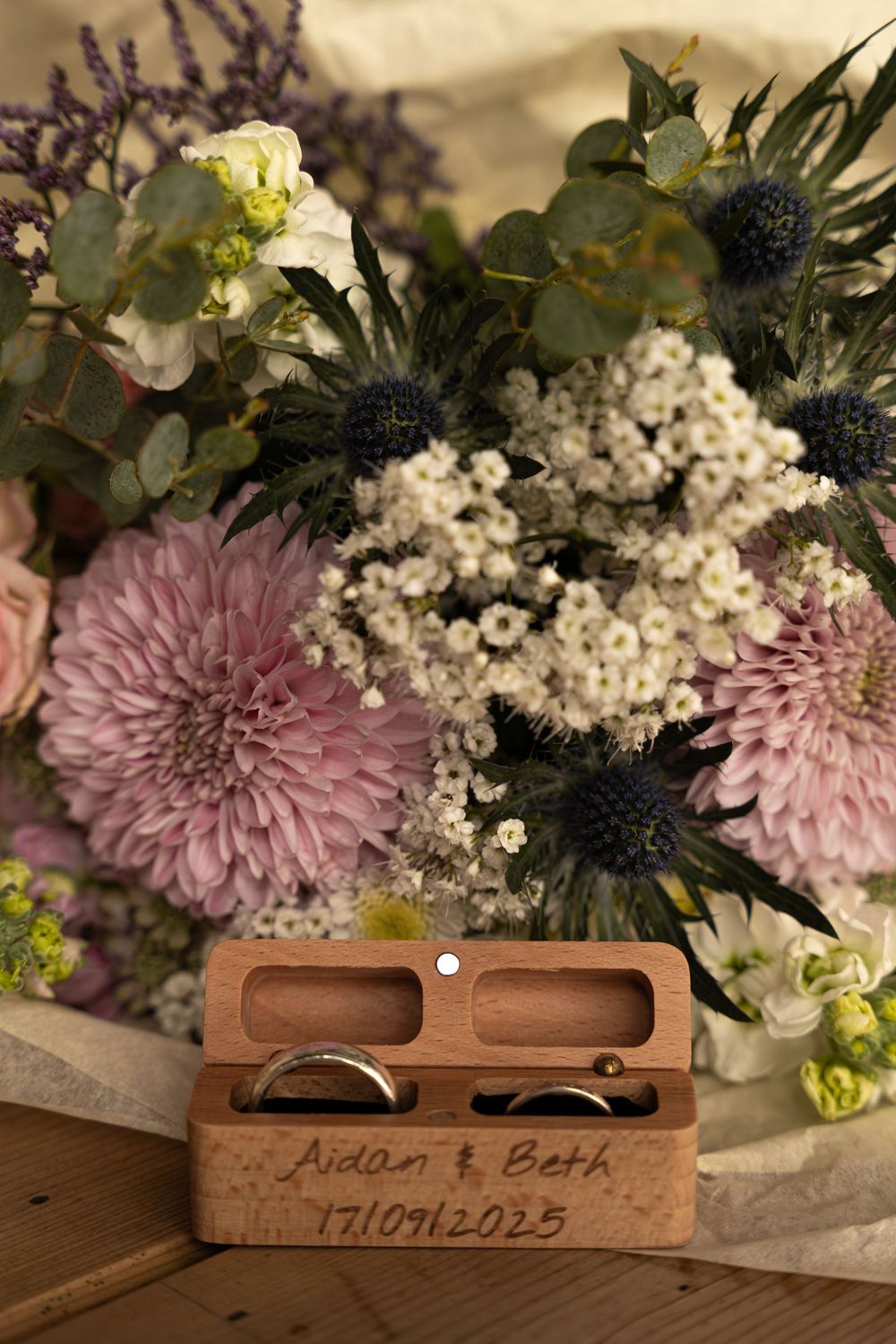Two wooden ring boxes with engraved date sit beneath a floral arrangement of pink chrysanthemums and baby's breath.